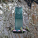 A teal glass bird feeder with a black tray and pink seed pieces, surrounded by four hummingbirds in a natural outdoor setting with blurred tree branches.