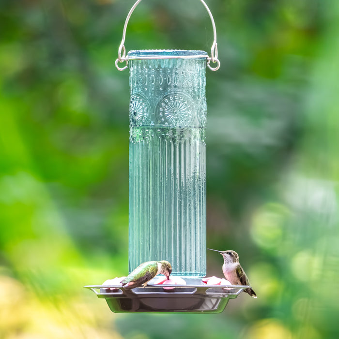 Decorative glass hummingbird feeder with two hummingbirds feeding on a blurred green background