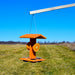 Orange bird feeder with a wooden platform hanging in an open field under a clear blue sky.