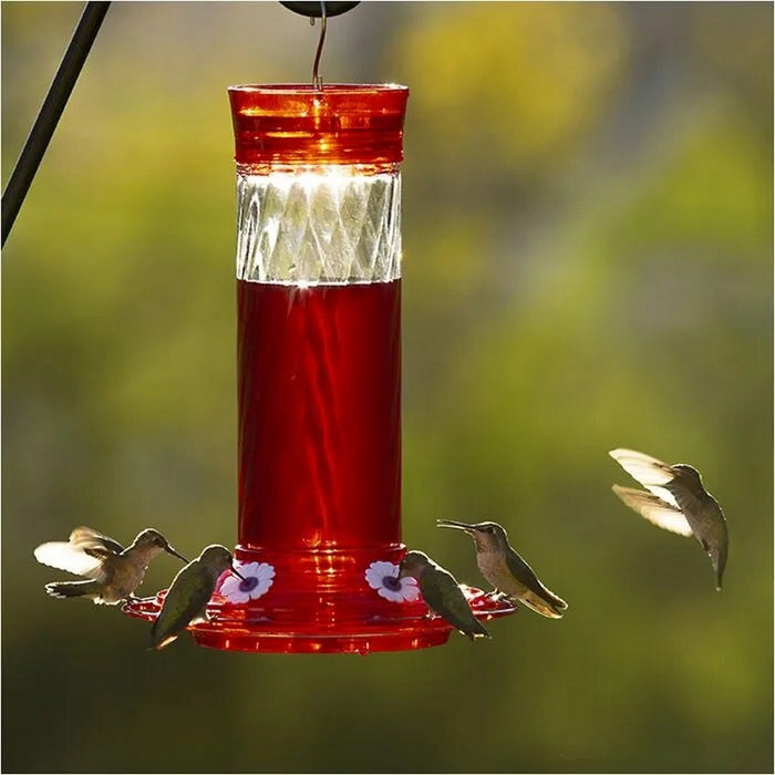 A red and clear hummingbird feeder filled with red liquid, shown outdoors with several hummingbirds around it against a blurred green and yellow background.