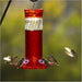 A red and clear hummingbird feeder filled with red liquid, shown outdoors with several hummingbirds around it against a blurred green and yellow background.