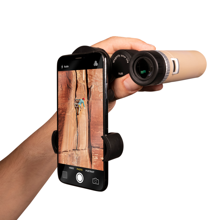 Hand holding a smartphone with a phone holder attached, displaying an image of a person climbing a rock wall. A telescope is pointed towards the screen.