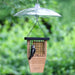 A wooden bird feeder with a clear dome-shaped rain cover and black hanging wires, shown outdoors with a small black-and-white bird perched on the feeder. The background is a soft-focus green garden.