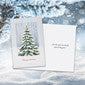 A Christmas greeting card featuring a green pine tree with white snow, shown next to a white envelope against a snowy outdoor background.