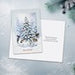 A square greeting card featuring a winter Christmas scene with a snow-covered pine tree, displayed on a light textured surface with a blurred festive background.