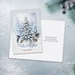 A square greeting card featuring a winter Christmas scene with a snow-covered pine tree, displayed on a light textured surface with a blurred festive background.