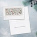 A close-up of a white greeting card with delicate pastel floral artwork at the top and a white lined writing area below, set against a soft light gray textured background.
