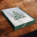 A square green gift box with a printed Christmas tree design on the lid, placed on a dark wooden surface.
