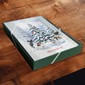 A square green gift box with a printed winter forest illustration and a red ribbon, placed on a dark wooden surface.