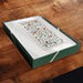 A square green gift box with a white floral patterned lid, placed on a dark wooden surface.