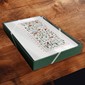 A square green gift box with a white floral patterned lid, placed on a dark wooden surface.