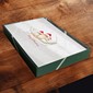 A square green gift box with a white printed logo and text, placed on a dark wooden surface.