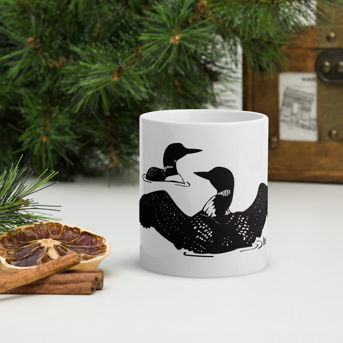 A white ceramic mug with a black loon illustration, styled on a white surface with a dried orange slice, cinnamon sticks, pine branches, and a wooden chest in the background.