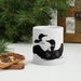 A white ceramic mug with a black loon illustration, styled on a white surface with a dried orange slice, cinnamon sticks, pine branches, and a wooden chest in the background.